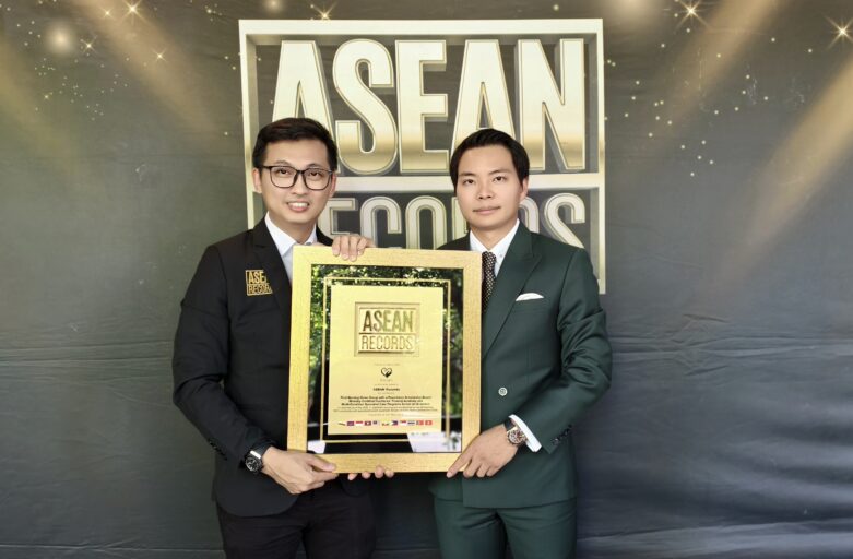Two men in formal suits pose together holding a gold-framed award plaque that reads ASEAN Records, with a large ASEAN-themed backdrop behind them.