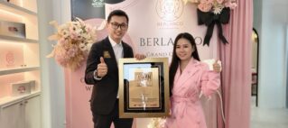 Man in a dark suit and a woman in a pink suit pose with a gold-framed award plaque, smiling at the camera, in a pink floral backdrop with BERLANCO branding behind them.