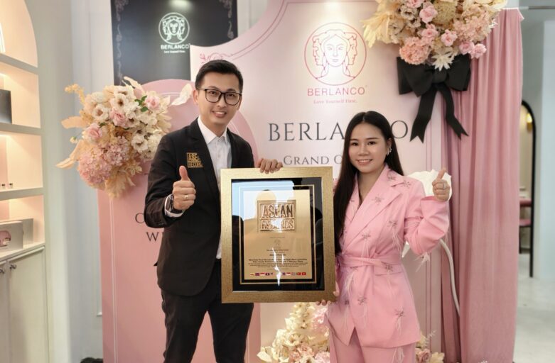 Man in a dark suit and a woman in a pink suit pose with a gold-framed award plaque, smiling at the camera, in a pink floral backdrop with BERLANCO branding behind them.