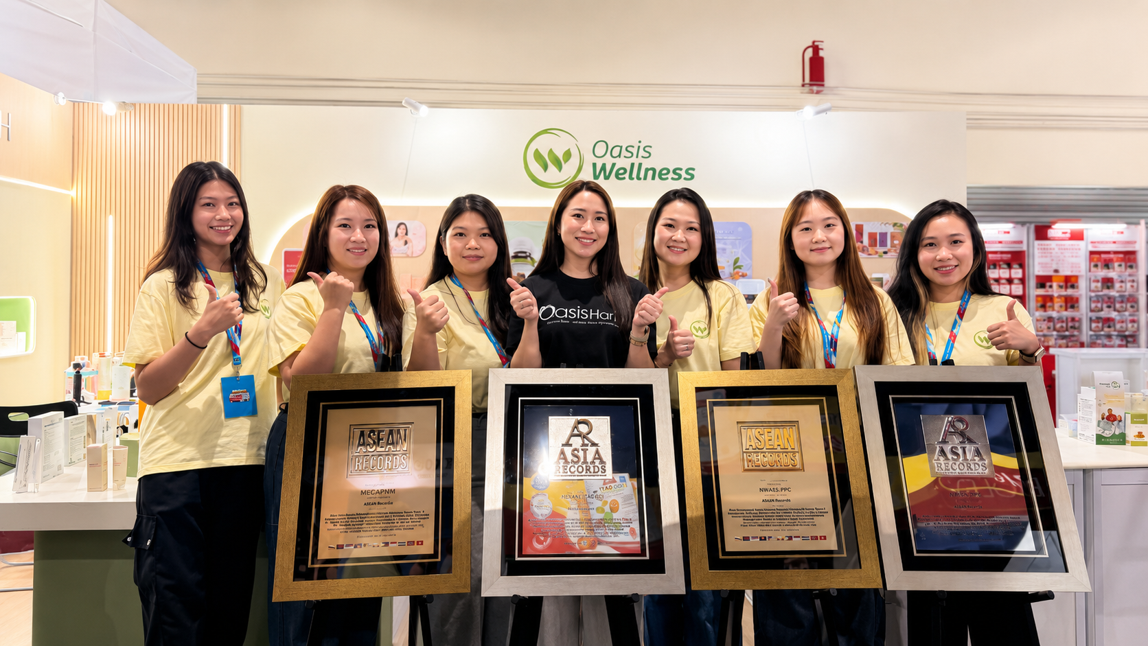 Seven staff members at the Oasis Wellness booth pose with four large framed awards in front of them, smiling and giving thumbs up.