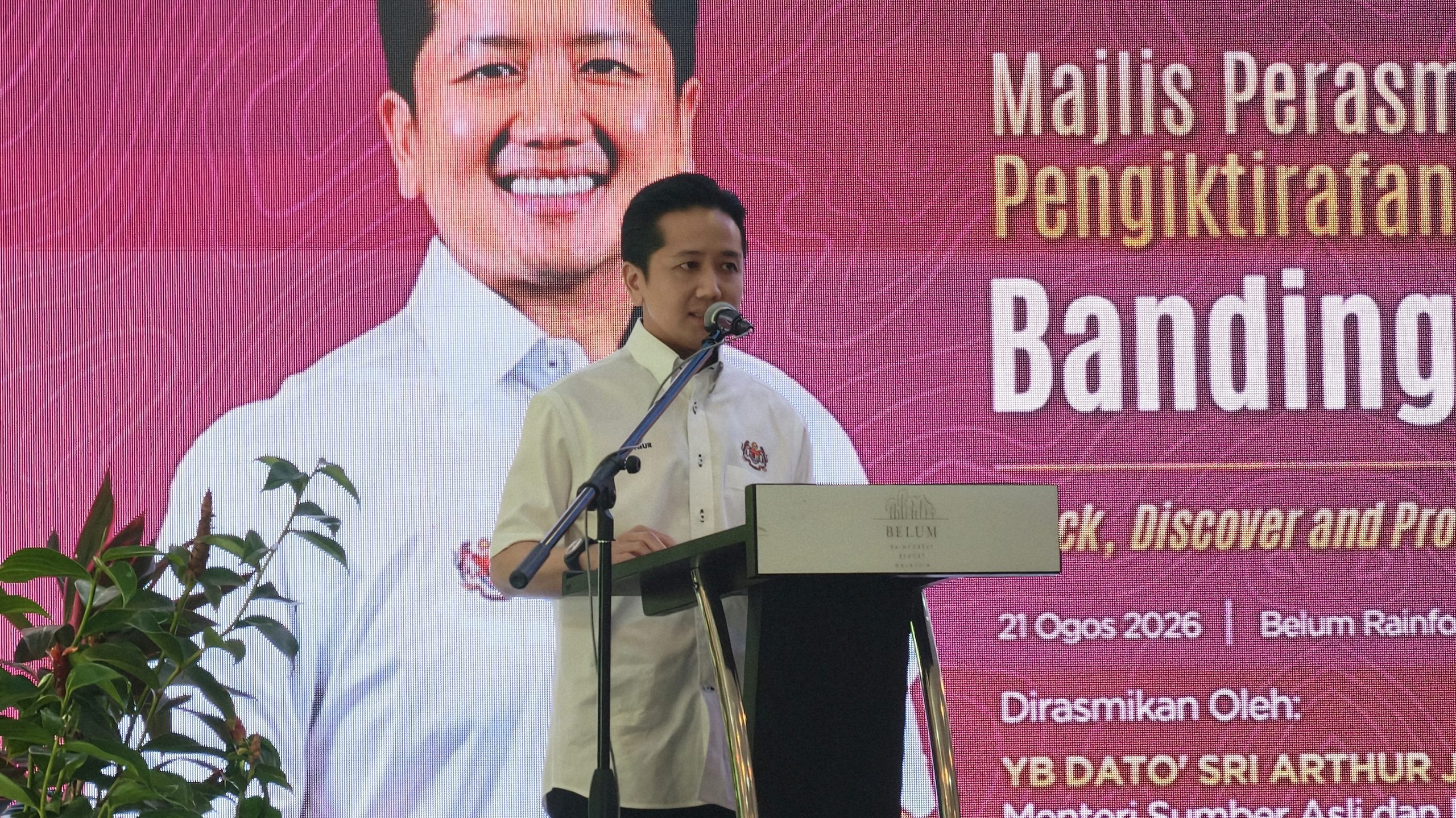 Man in light shirt speaks into a microphone at a podium, with a bright pink backdrop showing large text behind him and plants in the foreground.