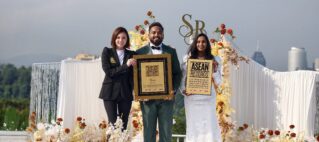 Three award recipients pose on a flower-decorated stage holding plaques, with a city skyline in the background.