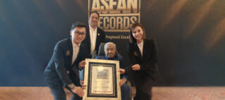 Elderly man in a wheelchair holding a framed award while three colleagues pose beside him in front of a backdrop that reads ASEAN Records.