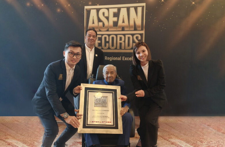 Elderly man in a wheelchair holding a framed award while three colleagues pose beside him in front of a backdrop that reads ASEAN Records.